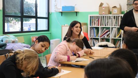 Infants asseguts i assegudes a unes cadires a una escola amb una noia donant-los suport.